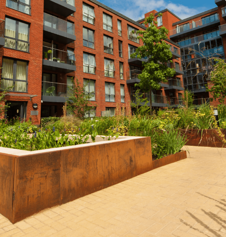 Exterior view of a modern red brick apartment building with large windows and balconies. In the foreground, lush green plants, bordered by elegant garden edging, thrive in raised flower beds along the paved sidewalk. The sky is clear and blue.
