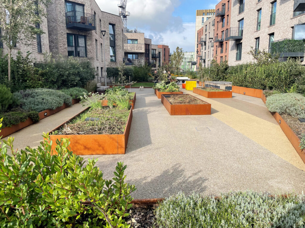 A modern apartment complex courtyard features CUSTOM raised garden beds, paved walkways, green shrubs, and surrounding multi-story buildings under a partly cloudy sky.