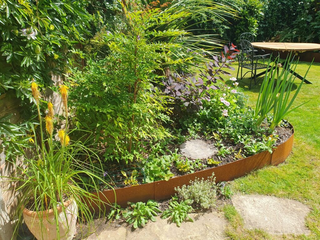 A sunny garden features a raised flower bed edged with rusty metal and filled with greenery, plus a round stone. A potted plant, grass, patio, and metal table with chairs appear in the distance. Shown: MINI TITAN (Pack of 2).