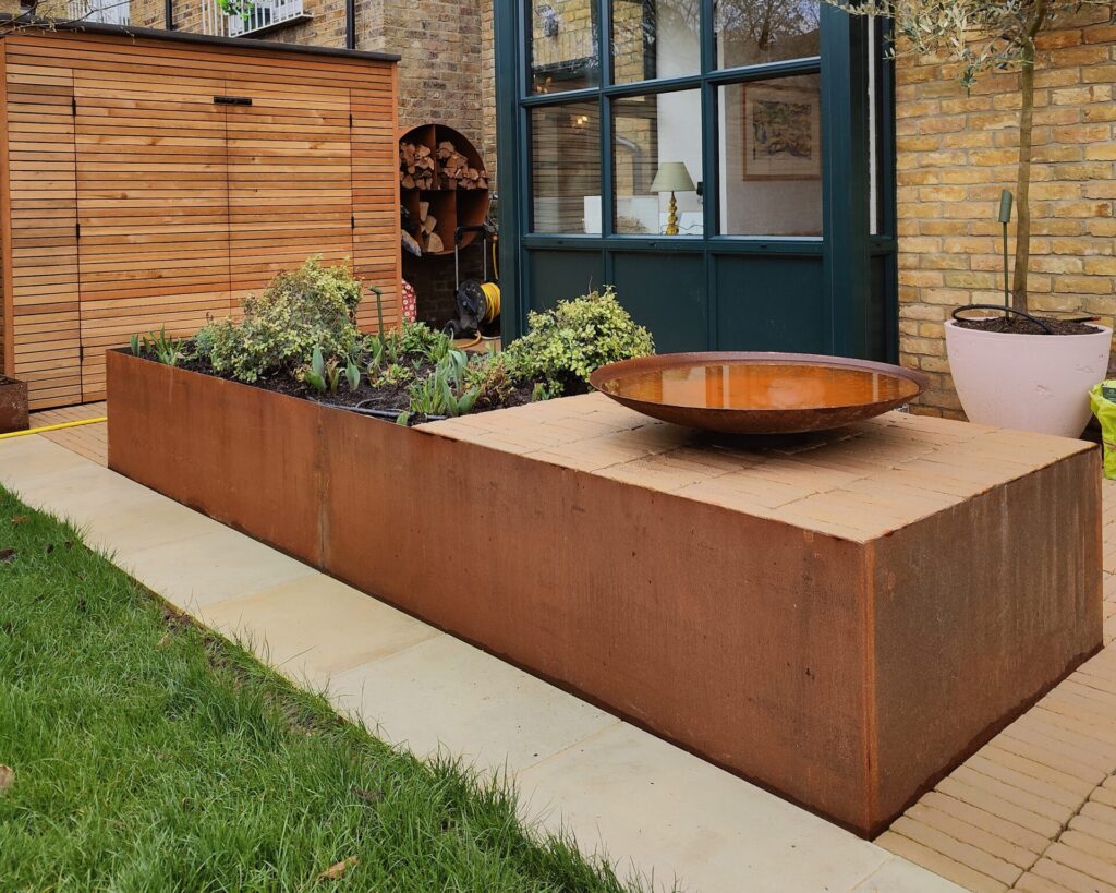 A modern garden showcases a large rectangular CUSTOM planter by EverEdge, filled with lush green plants and topped with a round metal water bowl. A wooden shed, potted plant, and brick wall complete this stylish setting.