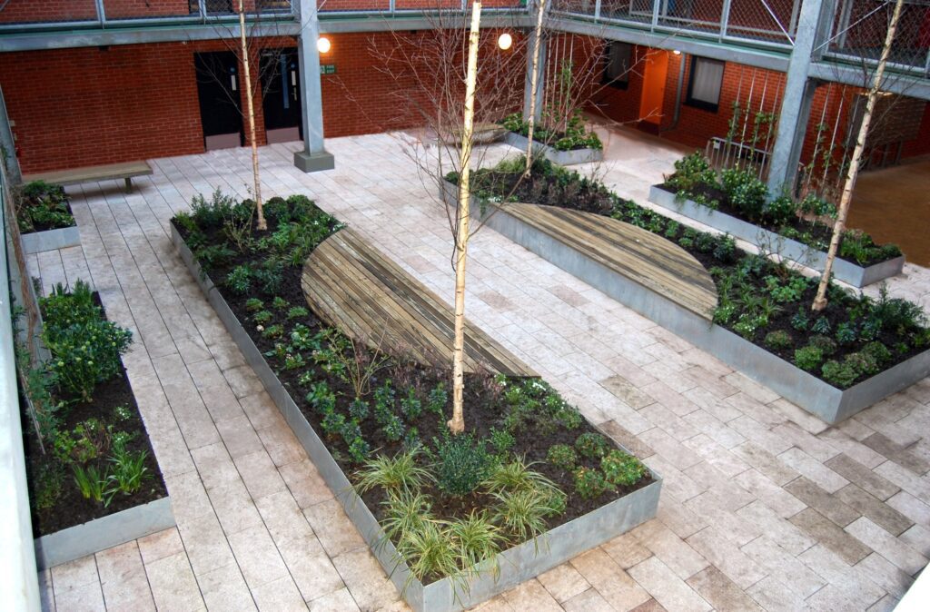 The modern courtyard features Custom Planter units filled with greenery, creating stylish garden borders. Light wooden benches blend seamlessly into the design, while a brick wall adds charm. Large rectangular tiles pave the area, completing this elegant outdoor space.
