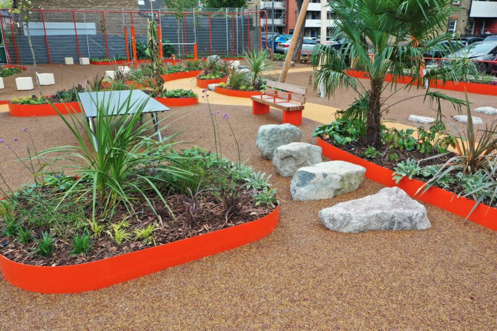 A modern urban garden features orange-edged raised beds made with Custom steel products, showcasing various plants, large rocks, benches, surrounding buildings, parked cars, and a ground covered in brown gravel.