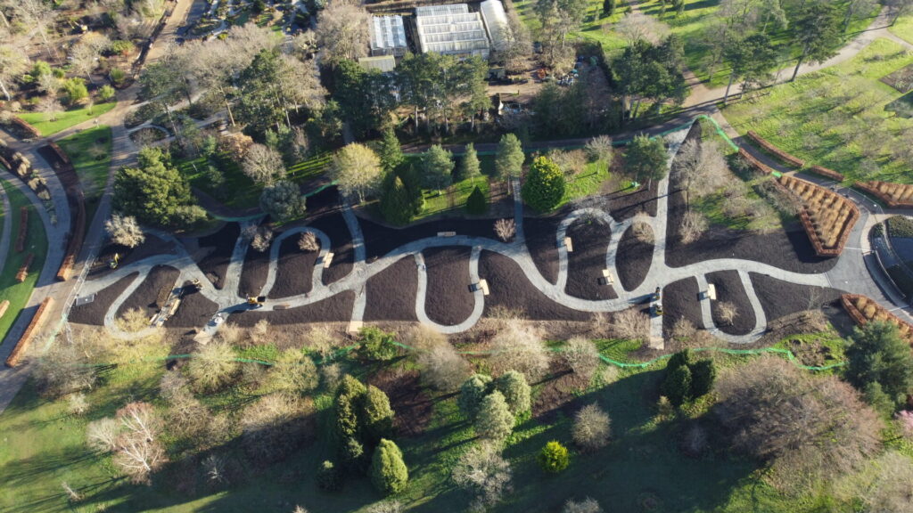 Aerial view of a winding, paved pathway bordered by TITAN heavy-duty steel edging in a landscaped park with trees, grassy fields, benches, and a greenhouse visible in the background.
