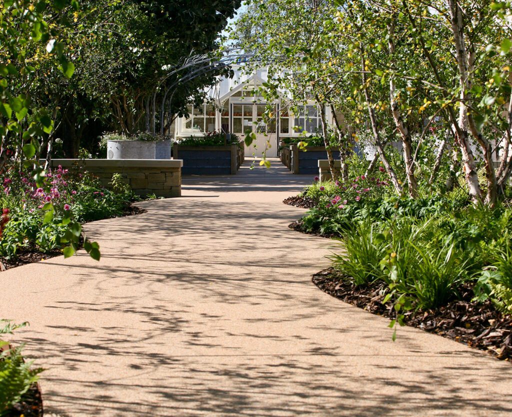 A winding path meanders through lush greenery, bordered by HALESTEM edgings of verdant plants and small trees. The path leads to a glass-walled building embraced by foliage under a clear sky, where branch shadows are elegantly cast along the way.