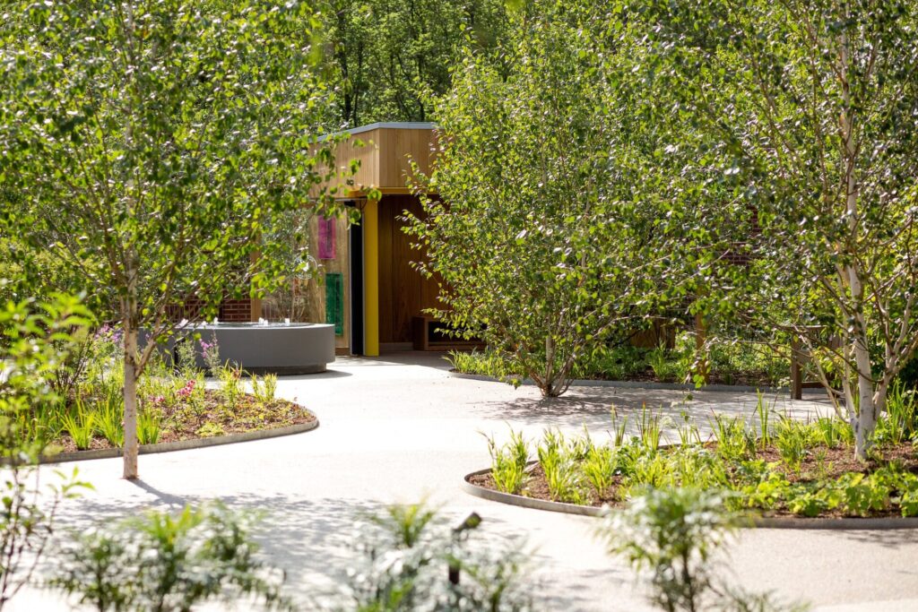 A winding, light-colored pathway bordered by TITAN steel edging curves through a lush garden with leafy trees, leading to a modern wooden structure partially hidden by foliage as sunlight brightens the vibrant scene.