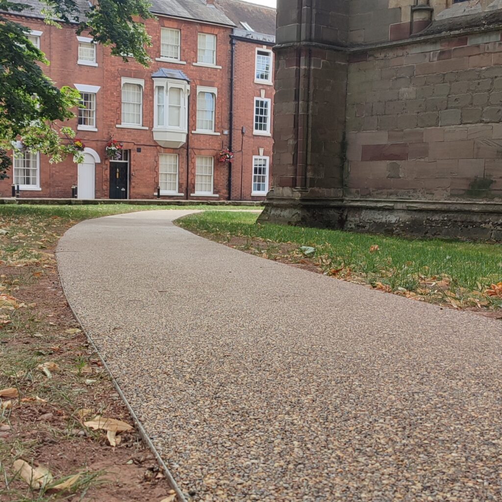 The HALESTEM features a curved gravel path next to a stone building, with red brick townhouses, green grass, trees, and scattered dry leaves in the background.