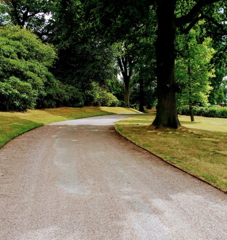 A winding wide gravel path curves through a green park with large trees and manicured lawns, creating a peaceful, shaded outdoor setting perfect for showcasing PROEDGE.