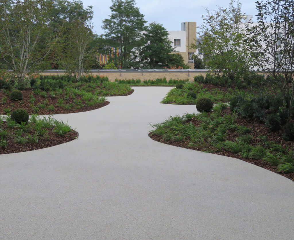 A curved concrete pathway, lined with HALESTEM garden edging, winds through neatly trimmed greenery, bushes, and small trees. Modern buildings rise in the background under a cloudy sky.