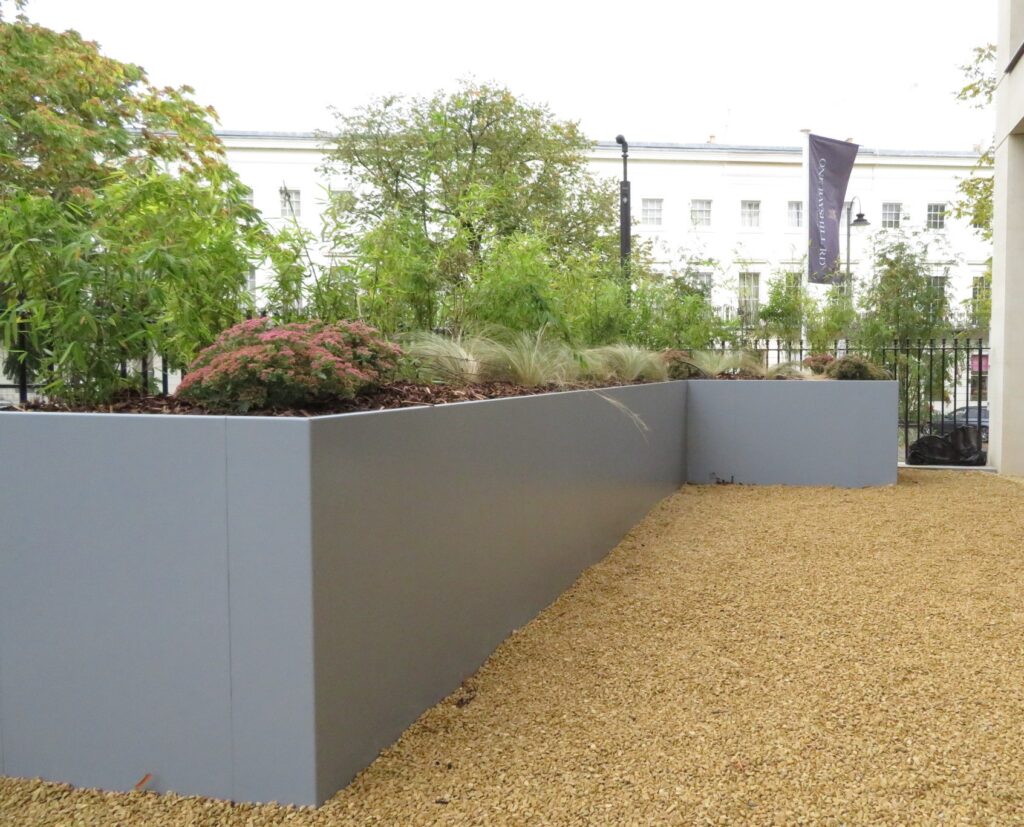 The EverEdge PLANTER, a modern grey raised planter filled with green plants and grasses, stands on light brown gravel with trees and a white building in the background.