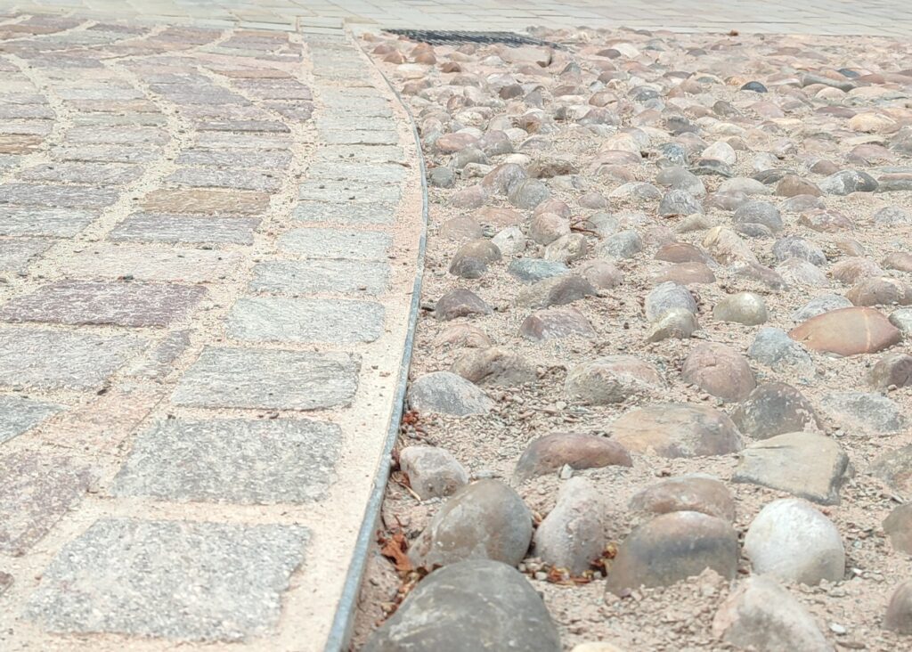 Close-up of a walkway featuring TITAN pavers on the left, creating a clear border with loose rocks and dirt on the right.