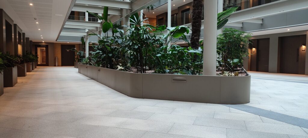 A modern indoor corridor with light tile floors and a large planter of lush green plants features CUSTOM by EverEdge, surrounded by doors and warm lighting.