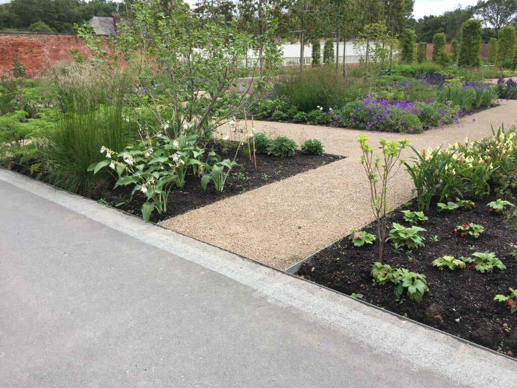 A HALESTEM garden features a winding gravel path, lush greenery with purple and white flowers, neat borders, trees, shrubs, a brick wall frame, and dense foliage under cloudy skies.
