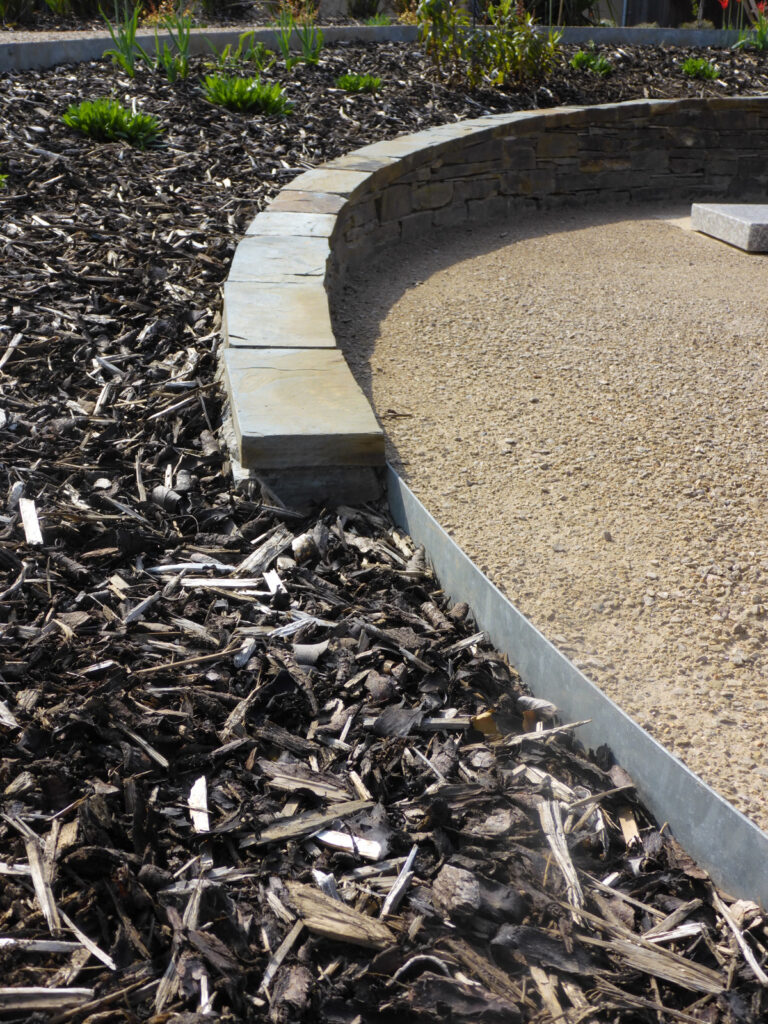 HALESTEM curved stone edging separates a mulched garden bed from a gravel path; sunlight illuminates green plants in the background.