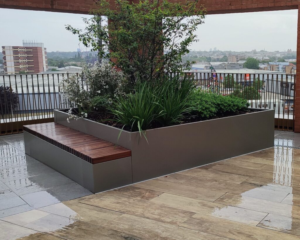 A modern rooftop terrace features the CUSTOM PLANTER—a large rectangular metal planter with green plants and small trees, plus an attached wooden bench—set on wet tiled flooring with city buildings in the background.