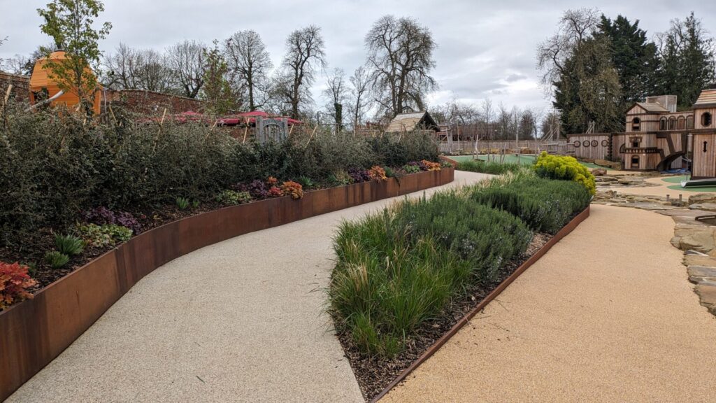 A winding pathway curves through a landscaped garden with raised flower beds bordered by CUSTOM steel edging, green shrubs, and wooden play structures beneath a cloudy sky.