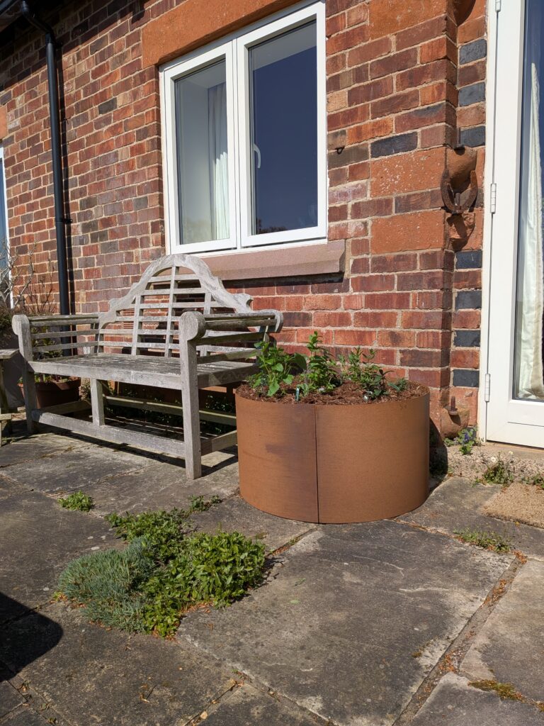 An EverEdge PLANTER filled with plants sits beside a weathered wooden bench on a stone patio by a brick house with white-framed windows and a glass door, while greenery grows between the patio stones.