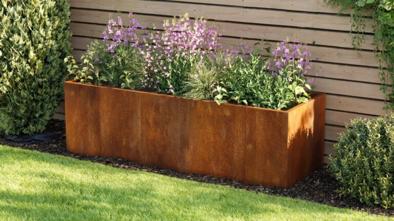 The EverEdge Planter, a rectangular rust-colored metal box, sits against a wooden fence filled with green plants and blooming purple flowers, surrounded by grass and shrubs.