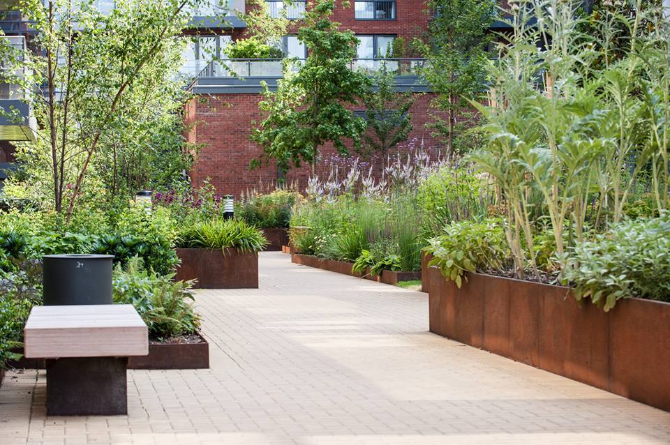 A landscaped courtyard features paved walkways, modern wood benches, lush trees, and raised rust-colored CUSTOM PLANTERs filled with greenery, all set against a red brick building.