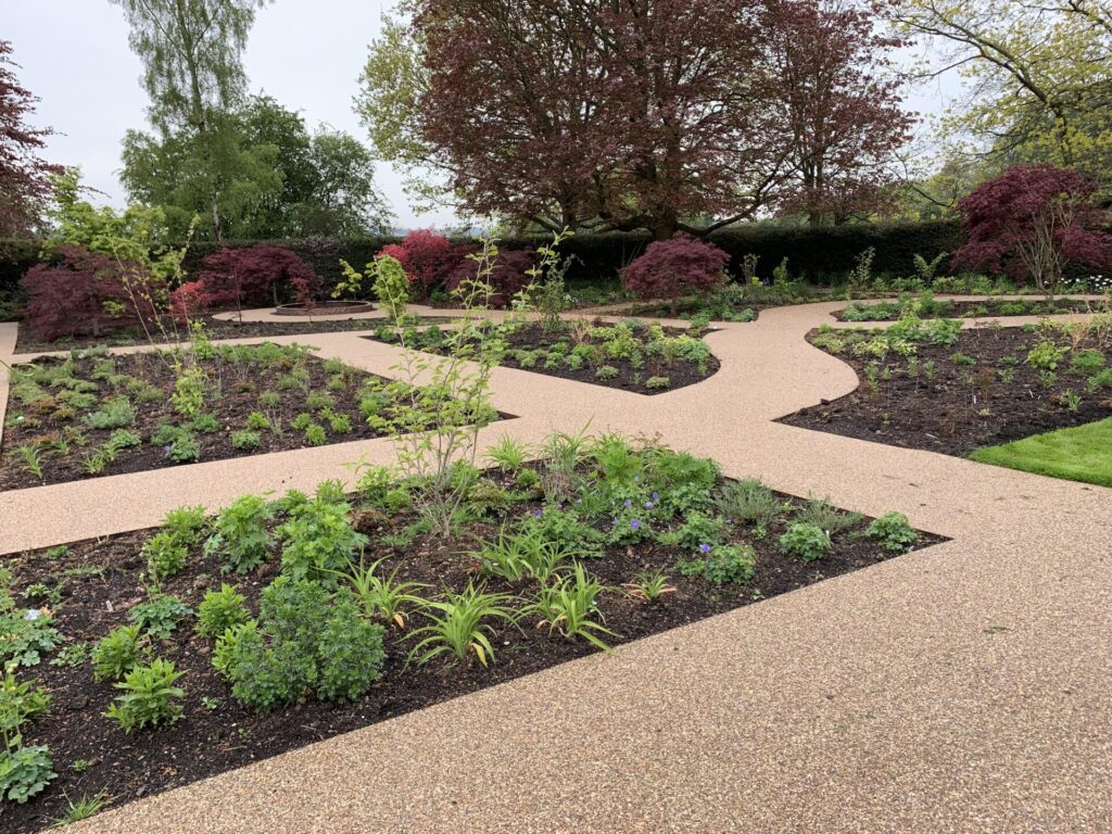 A landscaped garden features TITAN heavy-duty steel edging bordering gravel paths that weave among neatly planted flower beds, young green plants, and colorful bushes, all framed by trees and hedges on a cloudy day.