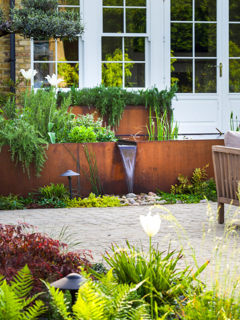 A modern garden features the CUSTOM CORTEN STEEL PLANTER in a rust finish, filled with tall green plants and white tulips. A small waterfall flows from the planter to a pond, with a wooden bench and a white door in the background.