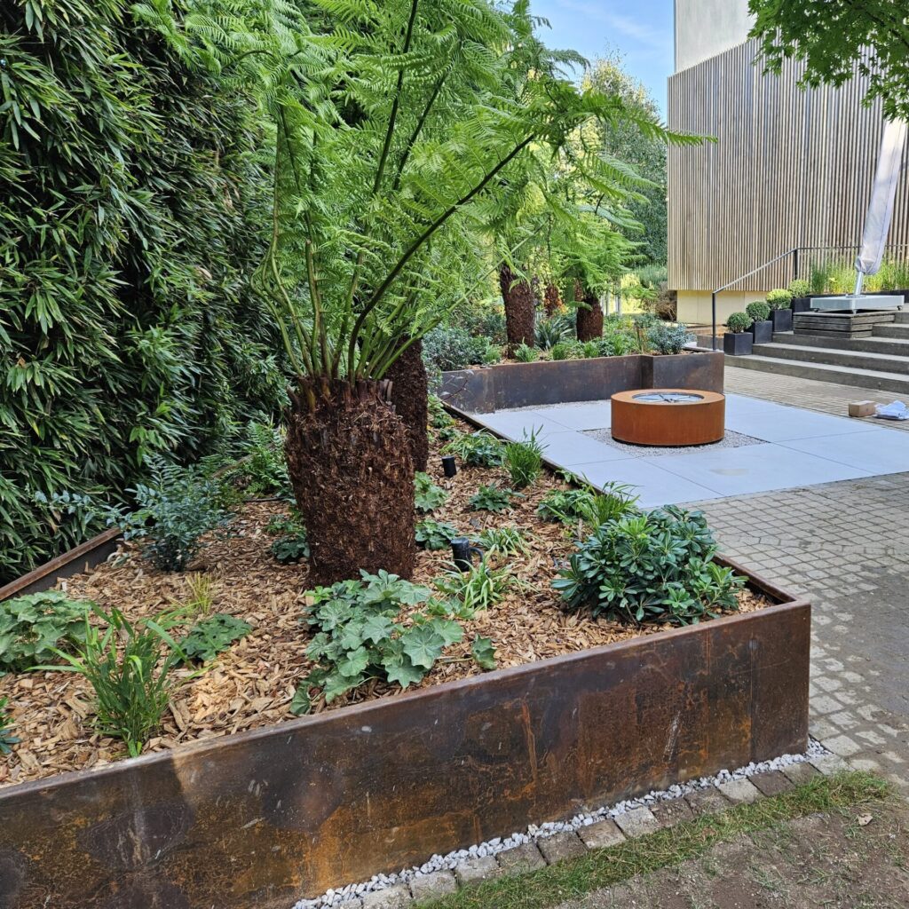 A modern garden showcases CUSTOM PLANTER beds with ferns and leafy plants, framed by rusted metal edging and wood mulch. A circular fire pit rests on a paved area with steps, set against a backdrop of lush greenery.