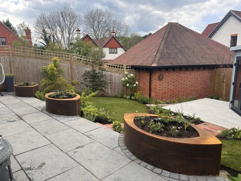 A landscaped backyard featuring CUSTOM PLANTER curved metal planters, young plants, a small lawn, wooden fencing, paving stones, and a red brick outbuilding with a tiled roof; trees and houses can be seen in the background.