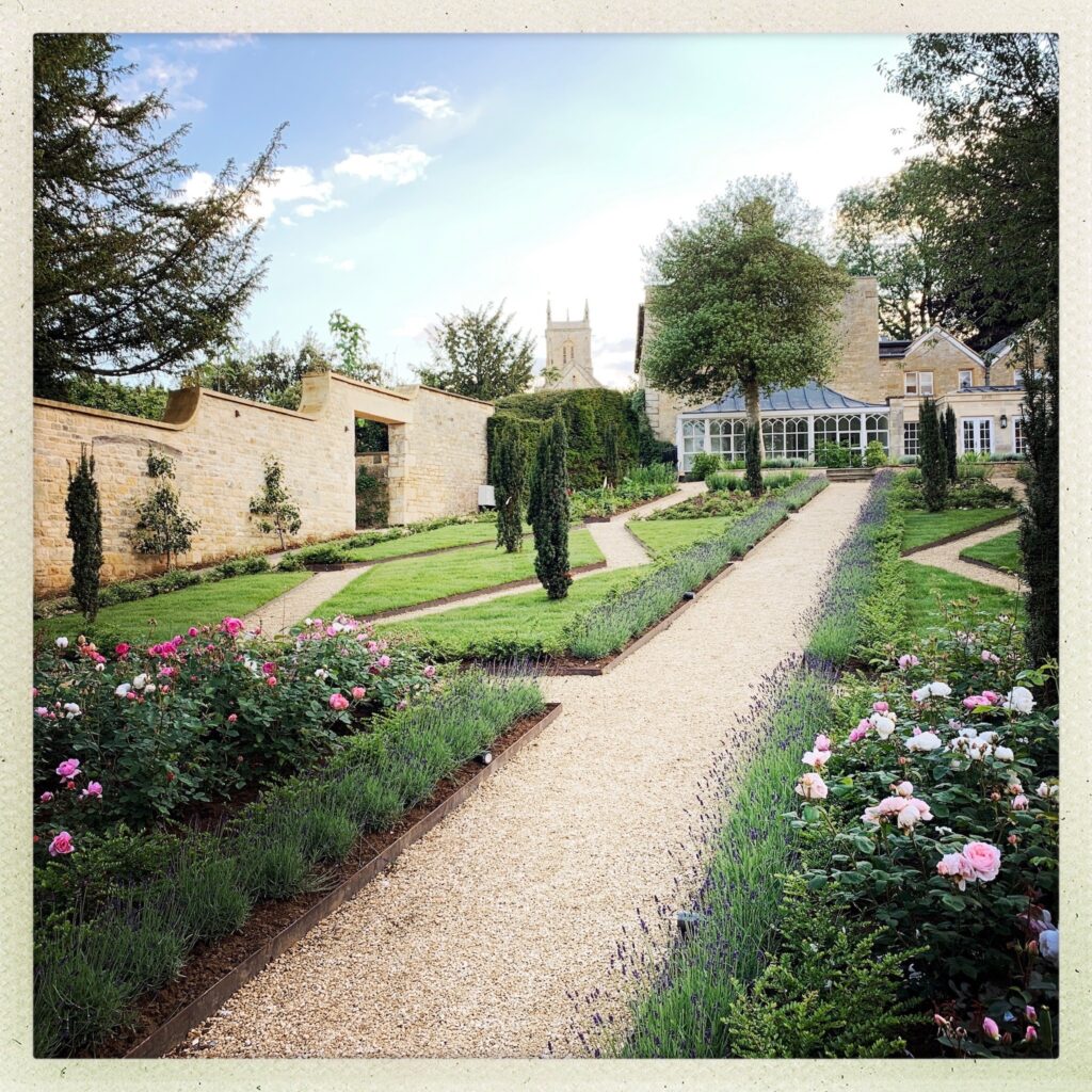 http://A%20picturesque%20garden%20with%20gravel%20paths,%20flowering%20lavender%20and%20roses,%20and%20manicured%20hedges%20defined%20by%20elegant%20garden%20edging.%20A%20stone%20building%20and%20church%20tower%20can%20be%20seen%20in%20the%20background%20under%20a%20partly%20cloudy%20sky.