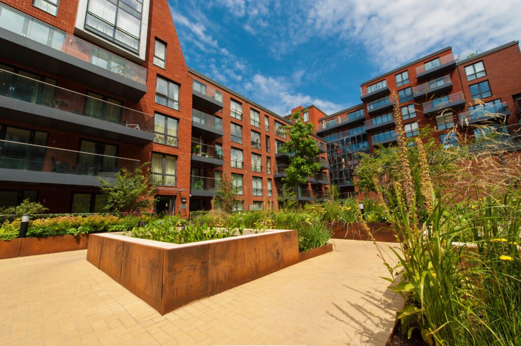 Modern apartment buildings with red brick facades and glass balconies surround a landscaped courtyard featuring garden borders and greenery, under a blue sky with scattered clouds.