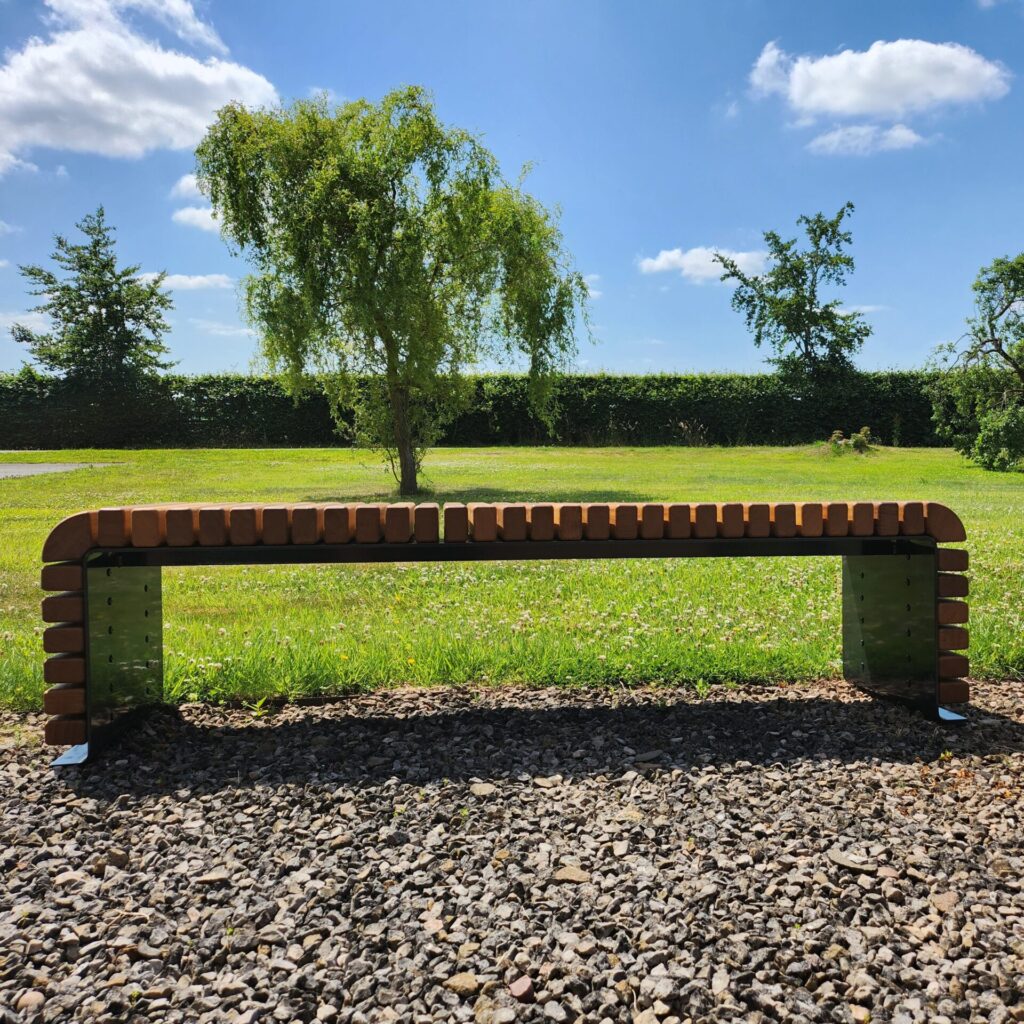 A STEEL BENCH sits on gravel in a sunny park, surrounded by green grass, leafy trees, and a blue sky with scattered clouds.