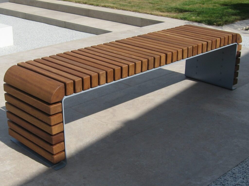 The STEEL BENCHES features a modern design with a wooden slatted seat and sides, metal legs, and is ideal for placement on stone patios near steps and grass.