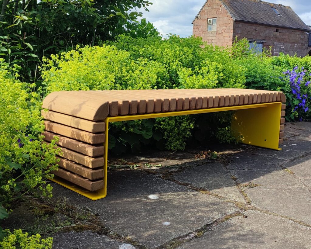 The STEEL BENCHES features a wooden slat top and sides with bright yellow metal legs. Placed on a stone patio among green bushes, it sits in front of a rustic brick building.