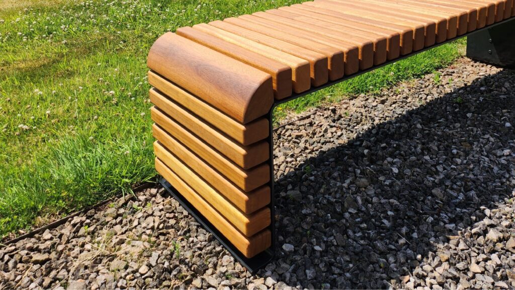 The STEEL BENCHES feature smooth wooden slats and a sturdy metal frame, positioned on gravel beside a grassy area in bright sunlight.