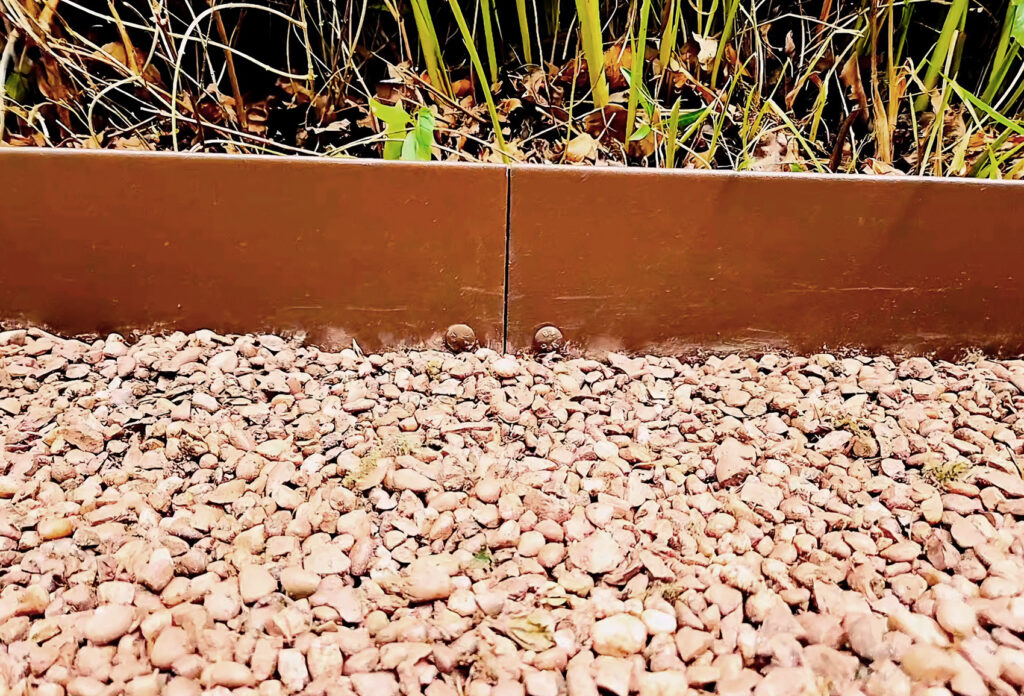 A close-up of the garden bed shows Rolltop - The Classic brown metal lawn edging, neatly separating a gravel path from verdant green plants. The gravel features a mix of small, rough stones in various gray and brown shades.