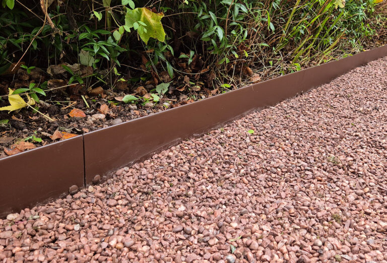 The ROLLTOP brown metal garden edging elegantly separates lush green plants from a path lined with reddish gravel, creating a clean, defined line between the verdant garden and rustic pathway.