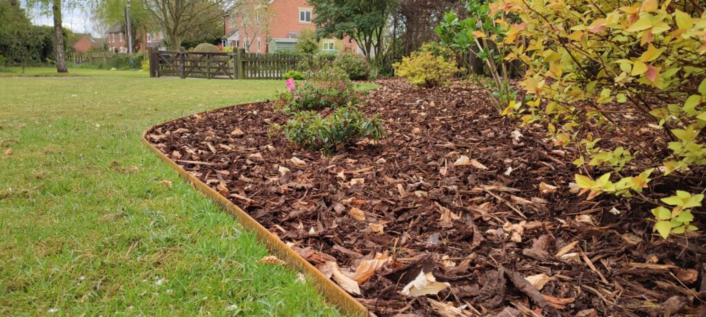 A garden bed lined with Rolltop - The Classic metal edging holds mulch and shrubs, bordered by green grass and trees, with residential houses visible in the background.