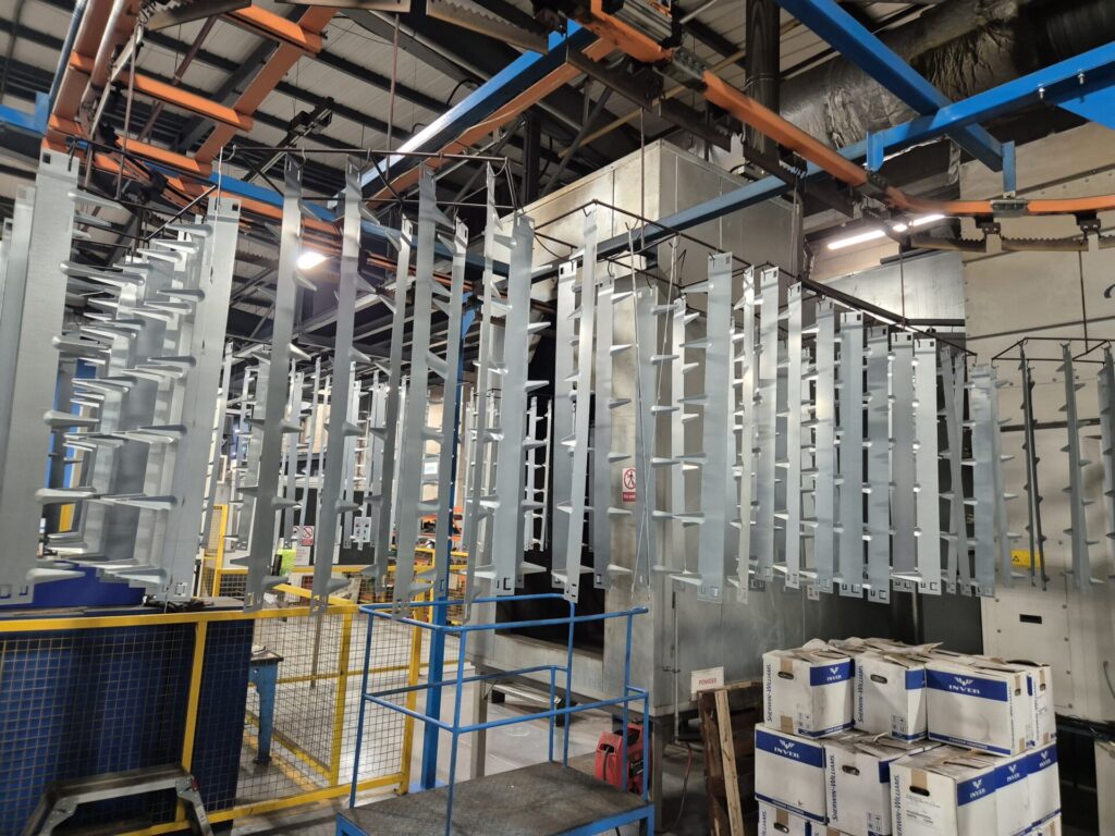 An industrial setting features metal structures hanging from conveyor tracks in a manufacturing facility. Boxes are stacked on the floor, and various pipes and machinery are visible in the background. Overhead lighting illuminates the scene, much like precise garden edging defining borders.