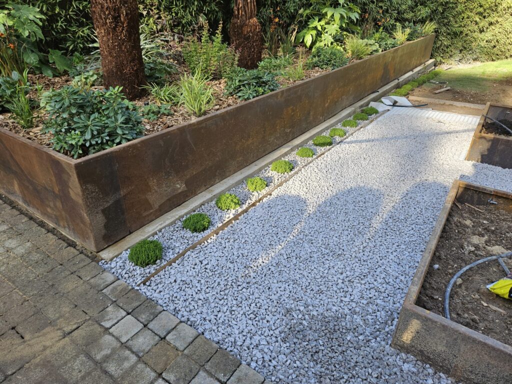 A landscaped garden features RETAINING WALLS (DRAFT), raised metal planters, a row of small green shrubs, and a pathway of light gray gravel bordered by flexible steel edging and paving stones. Sunlight casts tree shadows onto the path.