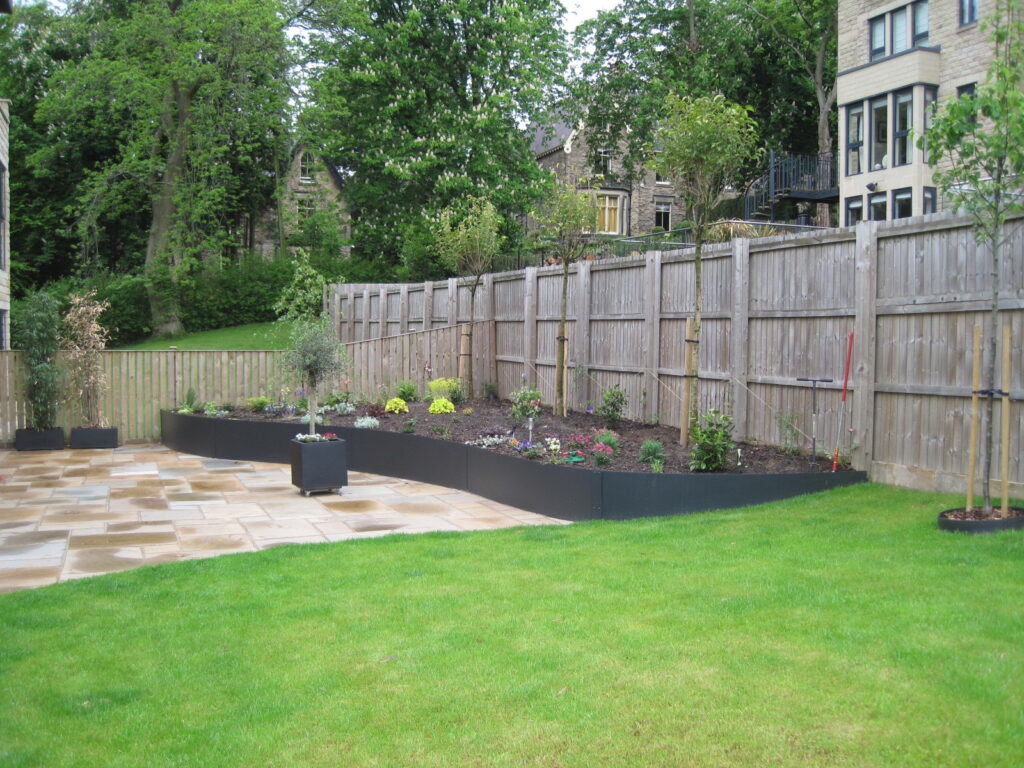 A backyard with a wooden fence, RETAINING WALLS outlining a curved raised flower bed filled with plants and flowers, a paved patio area, and a neatly mowed green lawn. Trees and houses can be seen in the background.