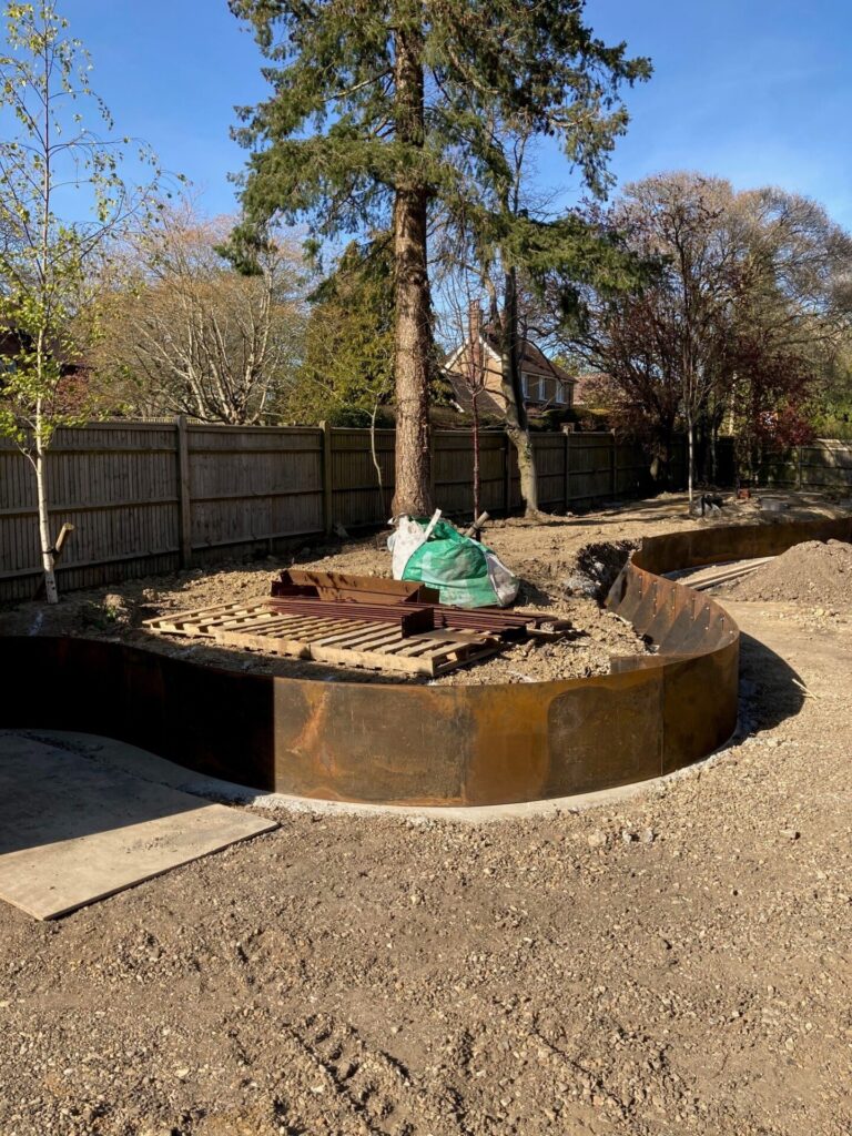A RETAINING WALLS (DRAFT) circular raised garden bed with flexible steel edging is being assembled on bare soil, with wooden planks and bagged materials on top; trees, a wooden fence, and houses are seen in the background.