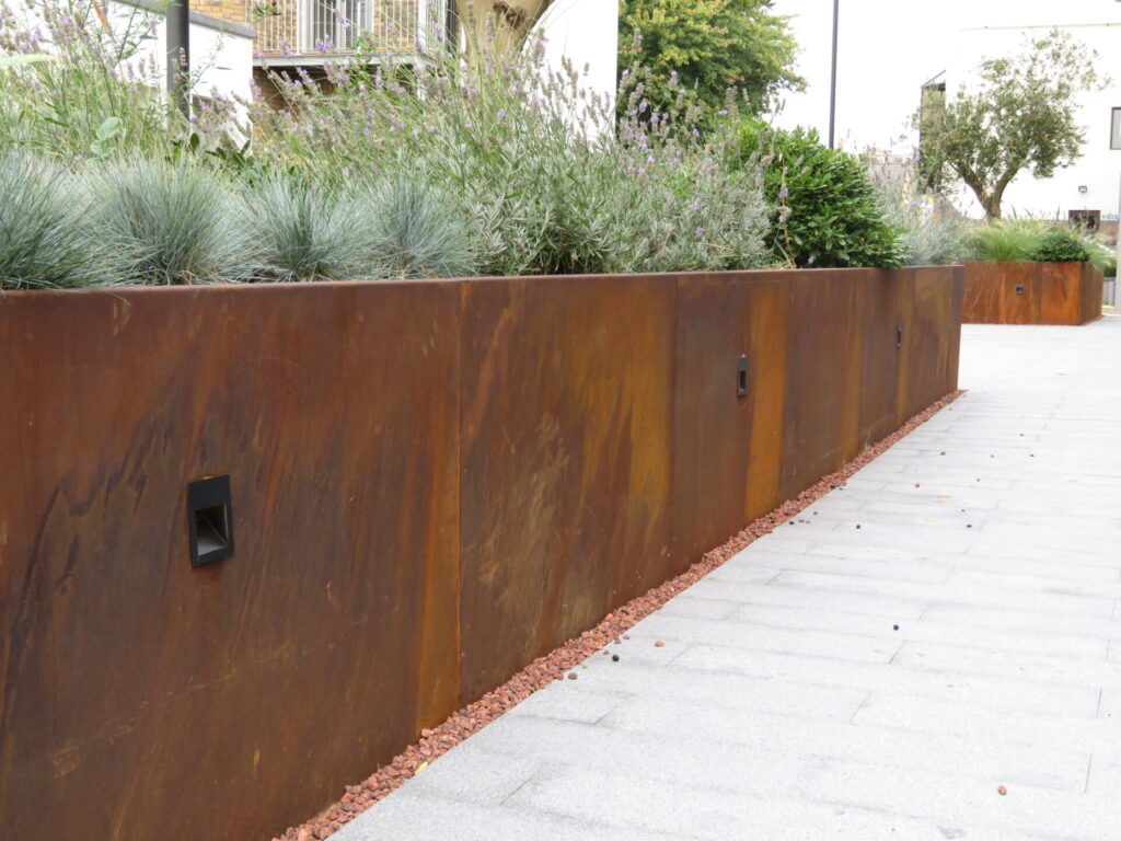 A rust-colored RETAINING WALL, accented by flexible steel garden edging, borders a path with ornamental grasses and shrubs. Small square lights are set into the wall, and the path is paved with light gray stone tiles.
