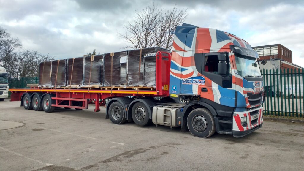 A large flatbed truck with a Union Jack design on the cab is parked, carrying several large, wrapped and secured items on its red trailer, in an outdoor industrial area with trees and a fence in the background.