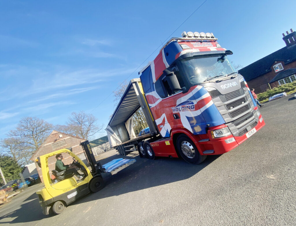 A forklift operator loads items onto a large truck decorated with a British flag design, parked on a sunny street near brick buildings and bare trees.