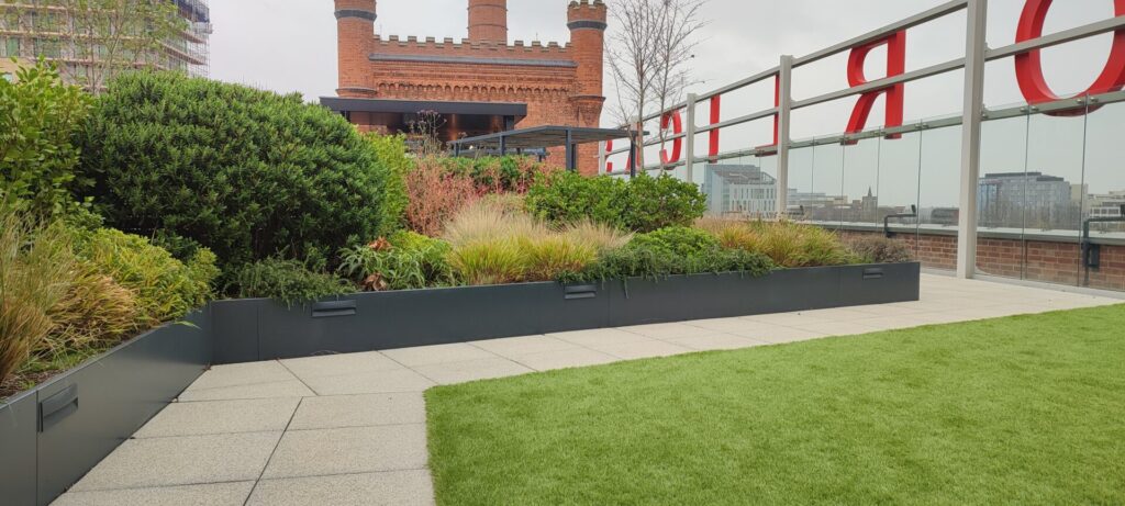 A rooftop garden features RETAINING WALLS, lush green plants, a glass wall with bold red letters, and a historic brick building with turrets. The ground is finished with paving stones and a patch of artificial grass.
