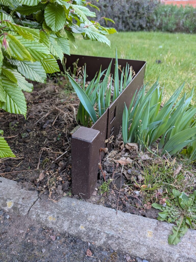 Finials brown metal garden edging separates a green plant bed from the lawn. The bed contains soil, trimmed leaves, and debris near a concrete curb, with shrub branches hanging over the left side.