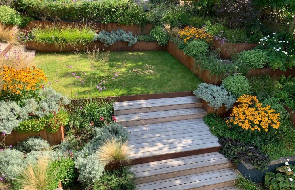 A vibrant backyard features terraced gardens with RETAINING WALLS (DRAFT), wooden steps, lush grass, flexible steel garden edging, and raised beds filled with yellow flowers, ornamental grasses, and green shrubs.