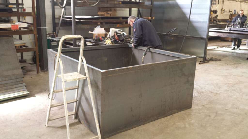 A person works on a large, rectangular metal container in a workshop. A small step ladder is positioned next to the container, and shelves with tools and materials are visible in the background.