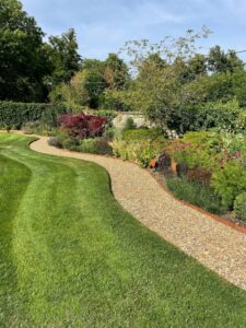 A winding gravel path runs through a neatly manicured green lawn, bordered by crisp lawn edging, colorful flowers, shrubs, and trees in a lush, sunlit garden.