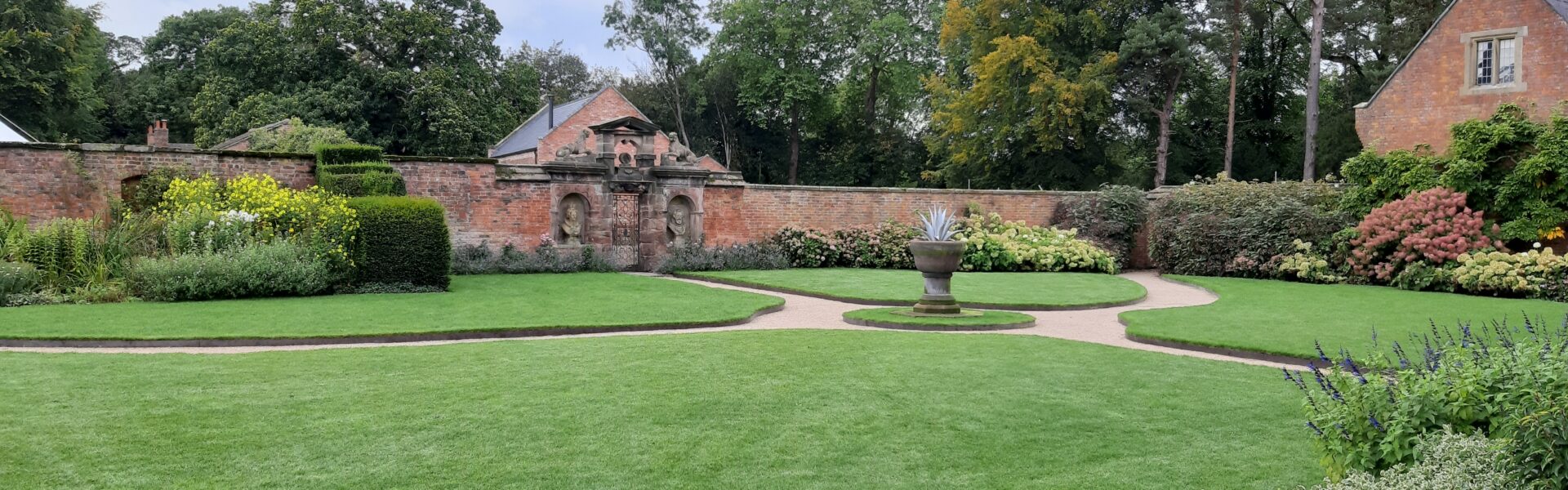 A well-kept garden with green lawns, colorful flowerbeds, a circular path, a large stone planter in the center, brick walls, and a red-brick building on the right under a cloudy sky.