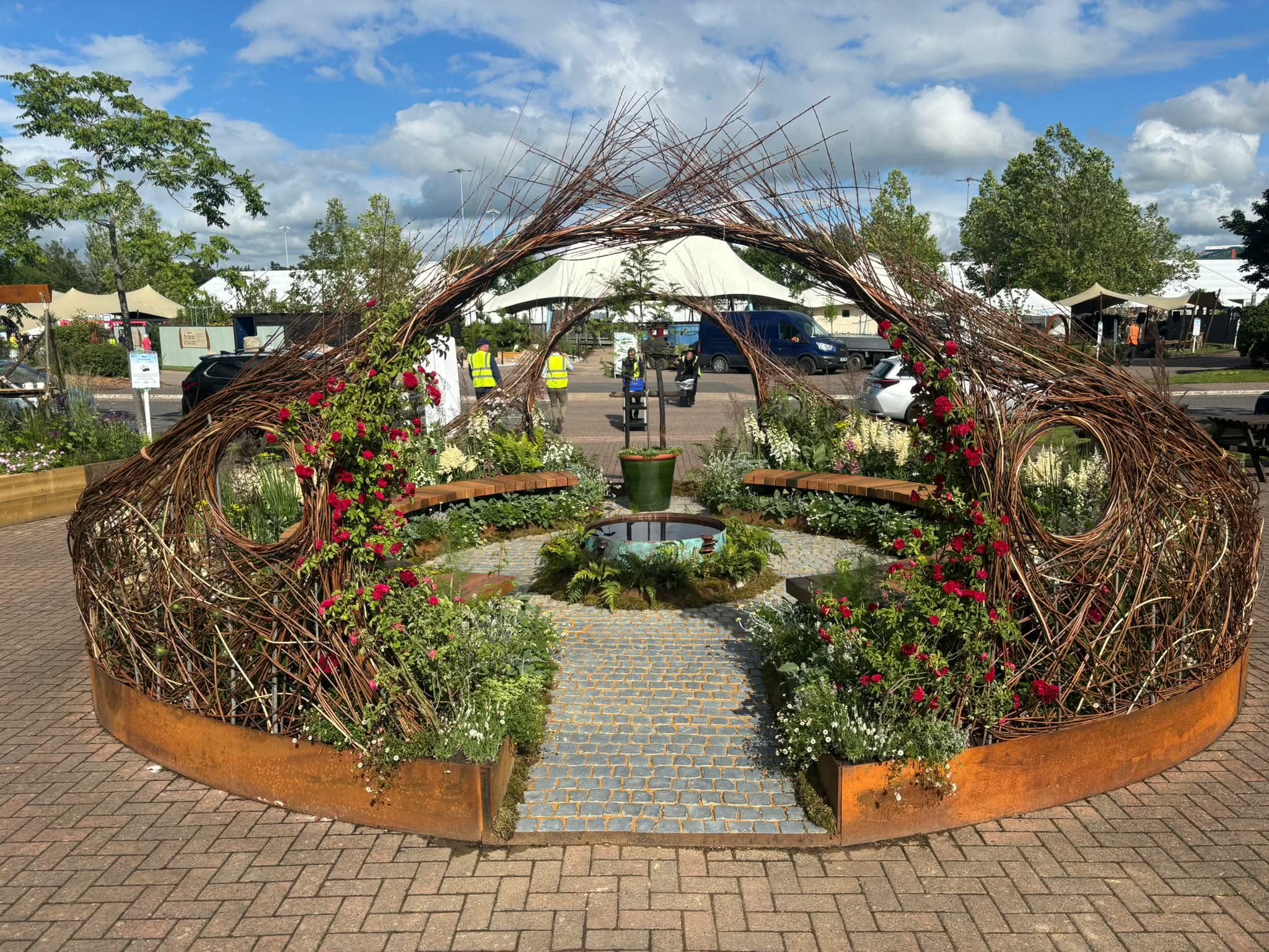 A circular garden structure made of woven branches and red flowers surrounds benches and a small central fountain on a paved area, with people and tents visible in the background.