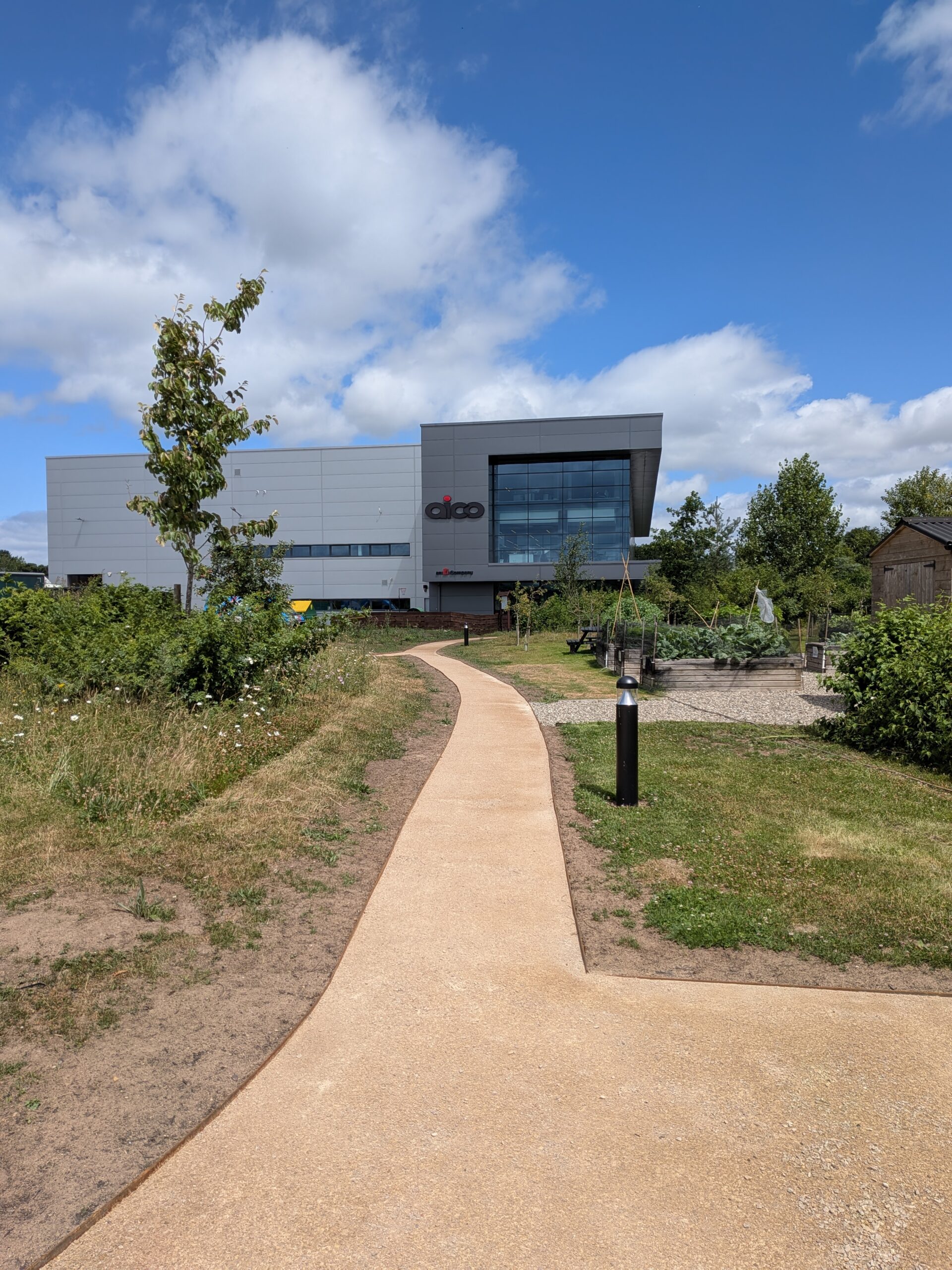A paved path leads through grass and greenery toward a modern, gray building with large windows under a blue sky with scattered clouds. There is playground equipment and trees on the right side.
